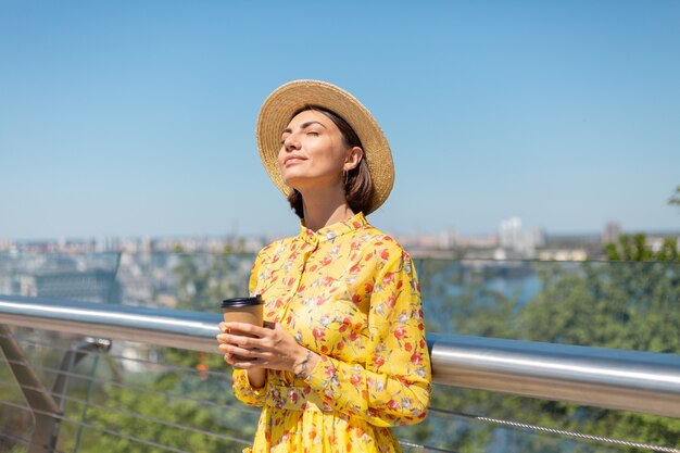 Retrato al aire libre de mujer con vestido amarillo de verano y sombrero con una taza de café disfrutando del sol, se encuentra en el puente con una vista increíble de la ciudad