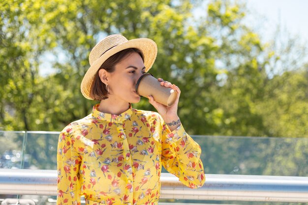 Retrato al aire libre de mujer en vestido amarillo de verano y sombrero con taza de café disfrutando del sol