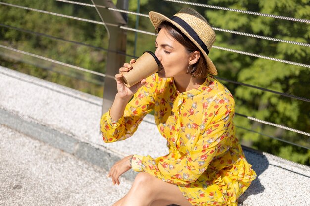 Retrato al aire libre de mujer en vestido amarillo de verano y sombrero con taza de café disfrutando del sol