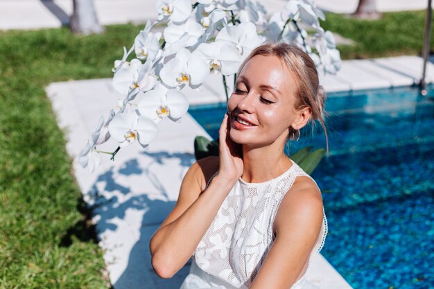 Retrato al aire libre de mujer en vestido de novia blanco sentada junto a la piscina azul con flores