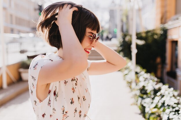 Retrato al aire libre de niña bonita riendo jugando con su pelo castaño corto en la calle. Foto de magnífica mujer caucásica divirtiéndose en verano.