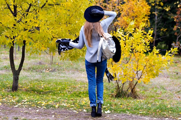 Retrato al aire libre de una niña emocionada viste de ala ancha de moda y de pie en pose de confianza. Mujer joven atractiva con gafas posando sobre fondo de naturaleza otoñal.