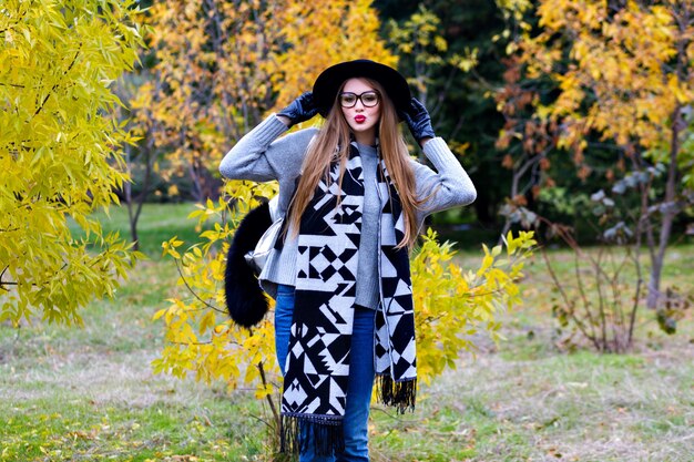 Retrato al aire libre de una niña emocionada viste de ala ancha de moda y de pie en pose de confianza. Mujer joven atractiva con gafas posando sobre fondo de naturaleza otoñal.