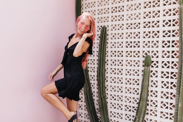 Retrato al aire libre de una niña encantadora lleva un vestido negro corto y gafas de sol bailando cerca de cactus.