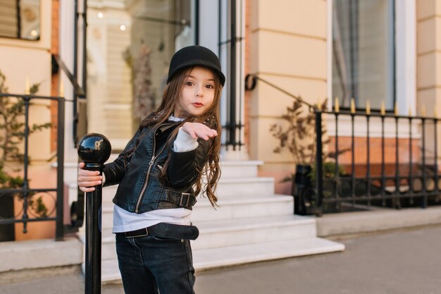 Retrato al aire libre de niña linda con sombrero enviando beso de aire con expresión de cara de sorpresa.