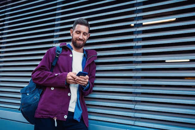 Foto gratuita retrato al aire libre de otoño o invierno de un guapo hombre hipster con barba, camiseta blanca, camisa azul y chaqueta marrón sosteniendo un teléfono inteligente.