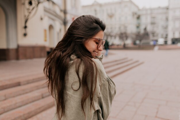 Retrato al aire libre de la parte posterior de la elegante mujer joven con chaqueta vestida de pelo largo disfrutando de tiempo fuera