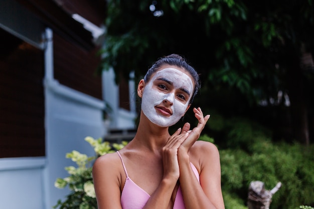 Retrato al aire libre de piel bronceada calma mujer bastante caucásica en bikini en el spa con mascarilla blanca en la cara