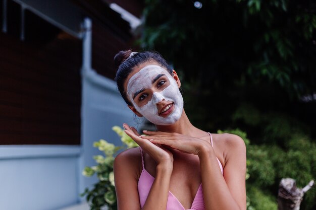 Retrato al aire libre de piel bronceada calma mujer bastante caucásica en bikini en el spa con mascarilla blanca en la cara