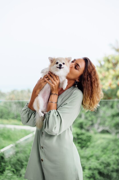 Retrato al aire libre de rizado mujer bronceada europea sostiene feliz perro mascota pomerania spitz