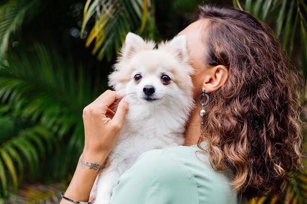 Retrato al aire libre de rizado mujer bronceada europea sostiene feliz perro mascota pomerania spitz