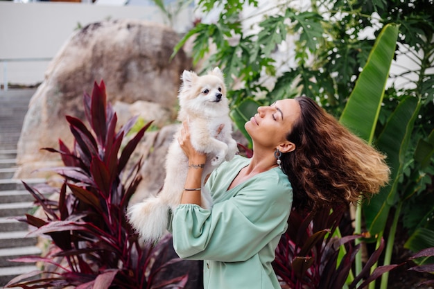 Foto gratuita retrato al aire libre de rizado mujer bronceada europea sostiene feliz perro mascota pomerania spitz