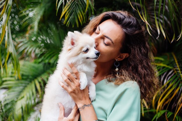 Retrato al aire libre de rizado mujer bronceada europea sostiene feliz perro mascota pomerania spitz