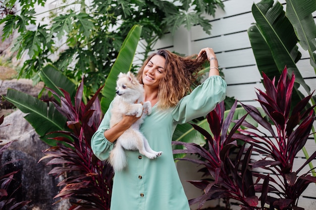 Retrato al aire libre de rizado mujer bronceada europea sostiene feliz perro mascota pomerania spitz