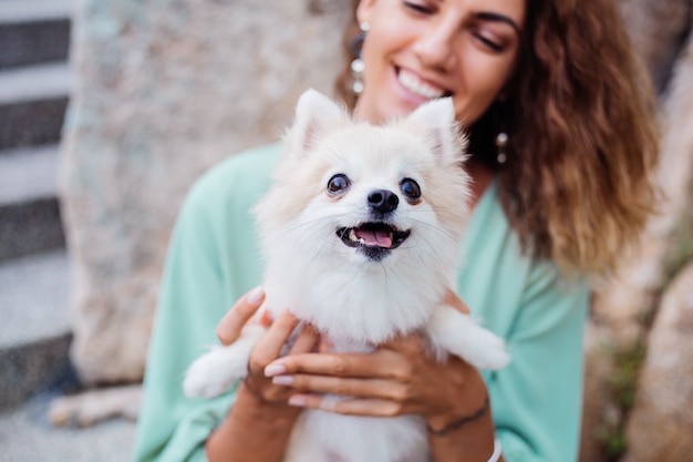 Retrato al aire libre de rizado mujer bronceada europea sostiene feliz perro mascota pomerania spitz