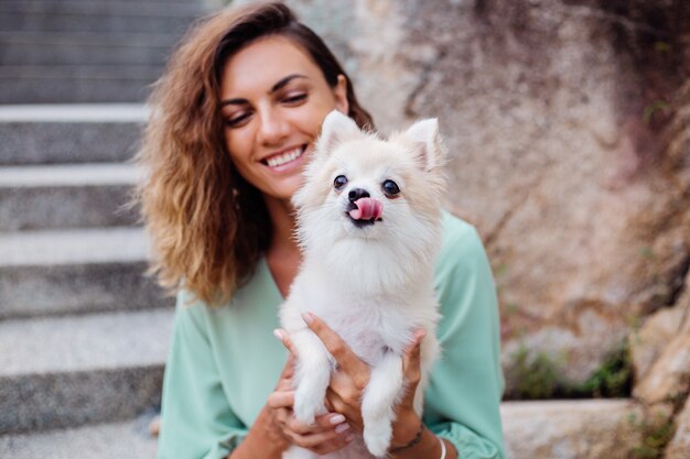 Retrato al aire libre de rizado mujer bronceada europea sostiene feliz perro mascota pomerania spitz