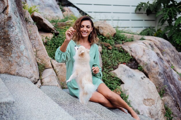 Retrato al aire libre de rizado mujer bronceada europea sostiene feliz perro mascota pomerania spitz