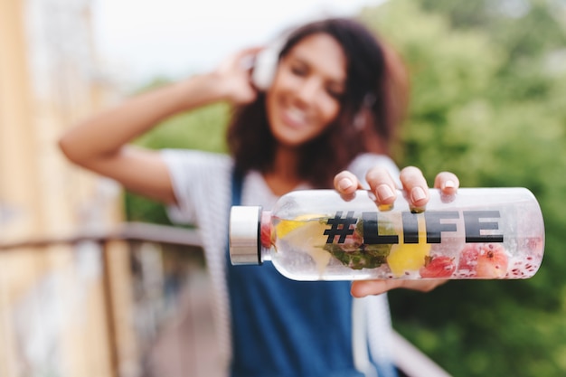 Foto gratuita retrato al aire libre de sonriente niña negra con camisa blanca con botella en la mano en primer plano