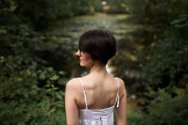 Retrato al aire libre de tierna joven mujer caucásica de pelo corto con piel pálida y cuerpo esbelto relajándose por estanque en el parque nacional