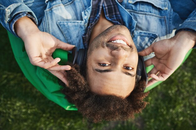 Retrato al revés de feliz joven deportista afroamericano acostado en el parque en la silla de bolsa de frijoles verdes, sonriendo a la cámara mientras sostiene los auriculares en los oídos y escucha música.