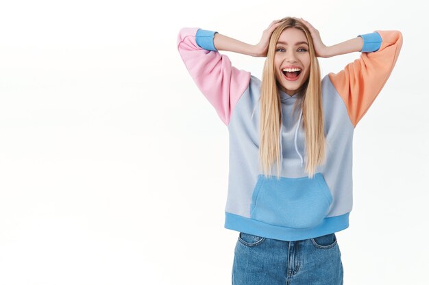 Retrato de alegre, afortunada chica femenina con cabello rubio, tomarse de las manos en la cabeza y sonriendo ampliamente, ganando