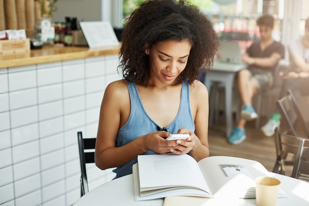 Retrato de una alegre y alegre mujer estudiante africana de piel oscura con cabello oscuro y rizado en camisa azul sentado en un café cerca de la universidad, leyendo un resumen académico, tomando café, conversando con su novio o