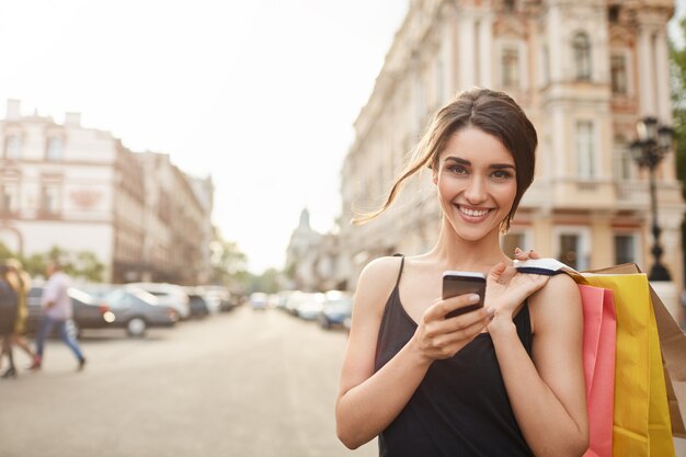 Retrato de alegre atractiva mujer caucásica joven con el pelo oscuro en vestido negro sonriendo a la cámara con los dientes, sosteniendo bolsas de la compra y el teléfono inteligente en las manos, catting con amigo. Enfoque suave