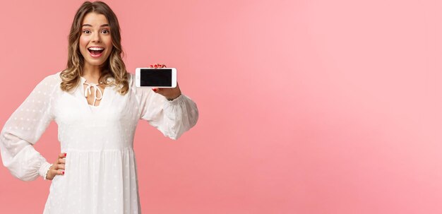 Retrato de una alegre y atractiva rubia caucásica vestida de blanco que muestra una pantalla de smartphone