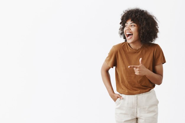 Retrato de alegre encantadora despreocupada mujer afroamericana con peinado rizado mirando y apuntando a la izquierda con pistola de dedo riendo de felicidad y buen humor