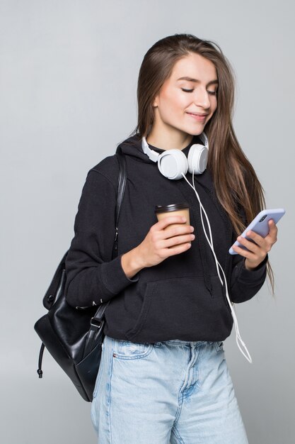 Retrato de una alegre estudiante atractiva con mochila escuchando música con auriculares mientras muestra la pantalla en blanco del teléfono móvil y baila aislado sobre la pared blanca