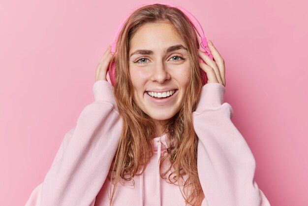 Retrato de una alegre y hermosa adolescente vestida con una sudadera informal que se pone las manos en los auriculares estéreo y escucha su música favorita mira felizmente directamente a la cámara aislada sobre un fondo rosa