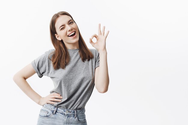 Retrato de alegre hermosa joven morena con longitudes de pelo medio en ropa casual elegante guiñando un ojo, con expresión satisfecha y alegre, mostrando gesto OK con la mano.