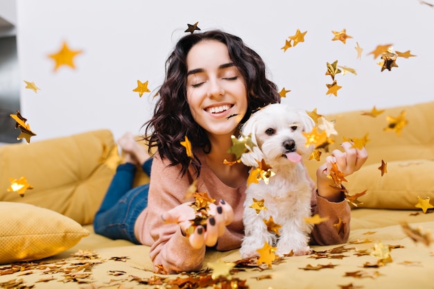 Retrato alegre increíble joven sonriendo con los ojos cerrados en la caída de oropel. Relajarse en el sofá con mascotas caseras, perrito blanco, sonriente, alegre, relajación