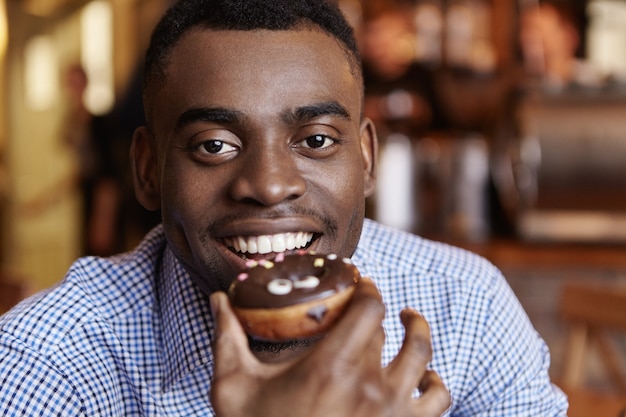 Retrato de alegre joven africano masculino vistiendo formal camisa a cuadros sosteniendo donut vidriado