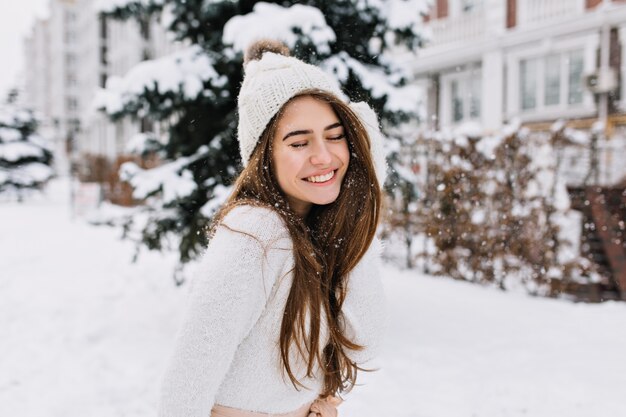 Retrato alegre joven con cabello largo morena divirtiéndose en la calle llena de nieve. Gorro de punto, suéter de lana blanca, sonrisa increíble, ojos cerrados, disfrutando del invierno.