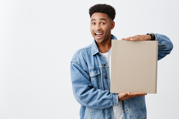 Retrato de alegre joven gracioso hombre de piel negra con peinado afro en camiseta blanca debajo de la chaqueta de mezclilla con cartón con expresión de la cara feliz y emocionado. Copia espacio