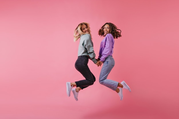 Retrato de alegre mejores amigos tomados de la mano en rosa. Hermanas encantadoras en pantalones de moda saltando y riendo.