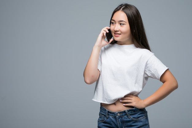 Retrato de una alegre mujer asiática hablando por teléfono móvil y mirando a otro lado sobre fondo gris