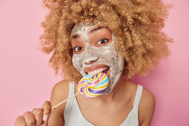 El retrato de una alegre mujer de cabello rizado muerde caramelo aplica una máscara de belleza en la cara para el tratamiento de la piel usa una camiseta casual aislada sobre fondo rosa Concepto de comida poco saludable