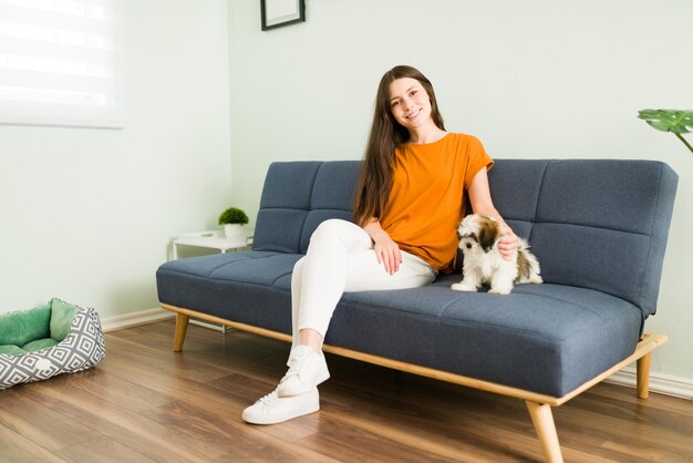 Retrato de una alegre mujer caucásica sentada en el sofá y disfrutando mientras pasa tiempo junto con su nuevo cachorro shih tzu