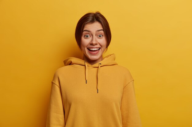 Retrato de una alegre niña milenaria ríe felizmente, escucha noticias agradables, usa una sudadera con capucha, tiene una conversación amistosa informal, una sonrisa blanca radiante, posa contra la pared amarilla,