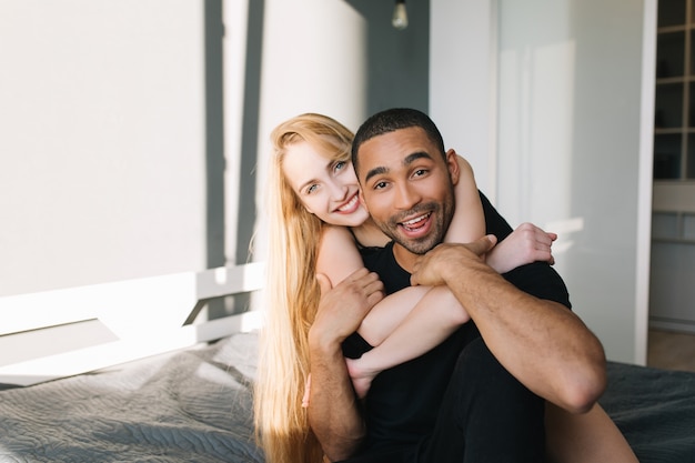 Retrato alegre pareja enamorada de linda joven con largo cabello rubio abrazando a chico guapo en la cama. Mañana soleada en casa, apartamento moderno, divirtiéndose, romance