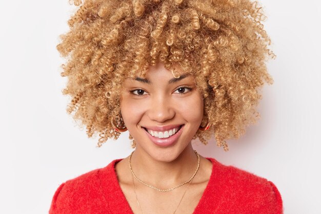 Retrato de alegre rubia rizada hermosa mujer sonríe felizmente muestra dientes perfectos incluso vestidos con poses casuales de puente rojo contra el fondo blanco del estudio Concepto de felicidad de las emociones de la gente