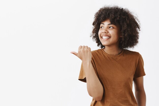 Retrato de alegre soñadora y despreocupada joven afroamericana con cabello rizado mirando y apuntando hacia la izquierda con el pulgar sonriendo de buenos recuerdos