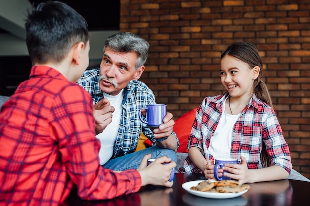 Retrato de alegre sonriente anciano bebiendo té con galletas mientras disfruta del tiempo con sus nietos.