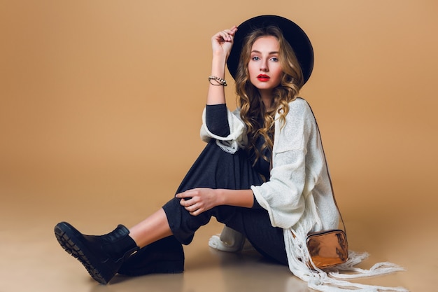 Retrato de alta moda de joven elegante mujer rubia con sombrero de lana negro con poncho de flecos blancos de gran tamaño con vestido largo gris.