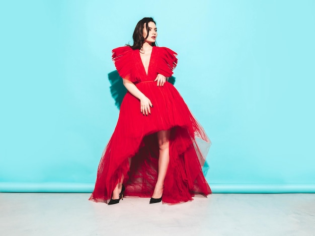 Retrato de alta moda de joven hermosa mujer morena con un bonito vestido rojo de noche esponjoso Modelo de moda sexy posando en el estudio Mujer de moda aislada en azul