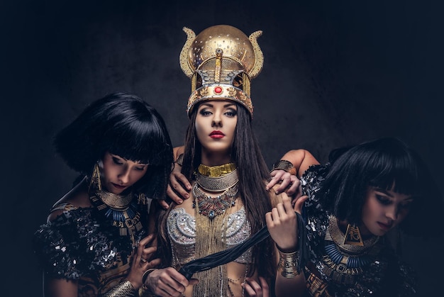 Retrato de la altiva reina egipcia con un antiguo traje de faraón con dos concubinas. Aislado en un fondo oscuro.