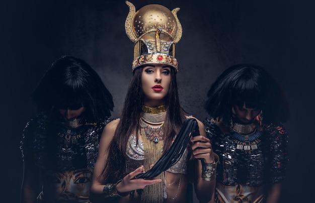 Retrato de la altiva reina egipcia con un antiguo traje de faraón con dos concubinas. Aislado en un fondo oscuro.