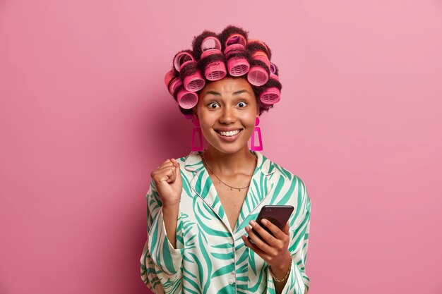 Retrato de ama de casa feliz aprieta el puño y sonríe ampliamente, vestida con una túnica doméstica informal, hace peinado, aplica rulos, espera una llamada, se regocija con las noticias positivas aisladas en la pared rosa